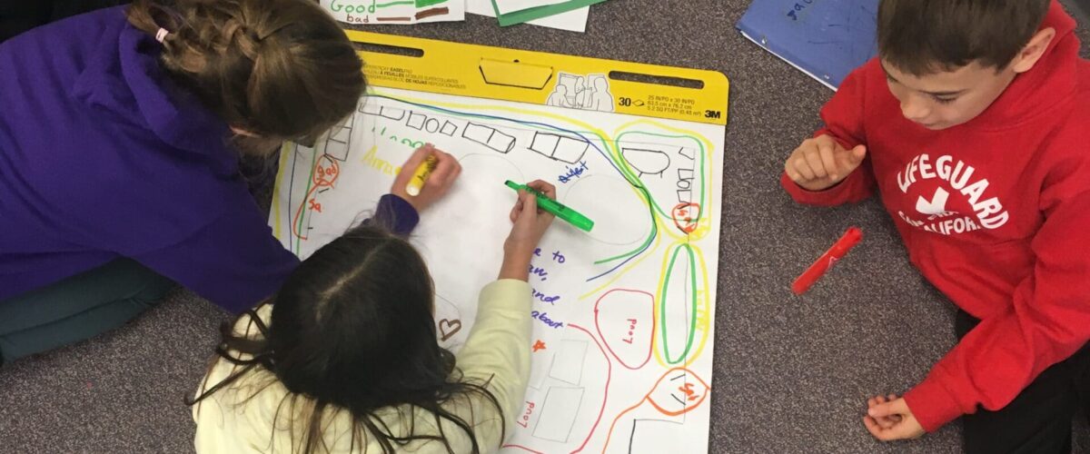 students sitting around a large writing pad with a drawing of a cafeteria on it.