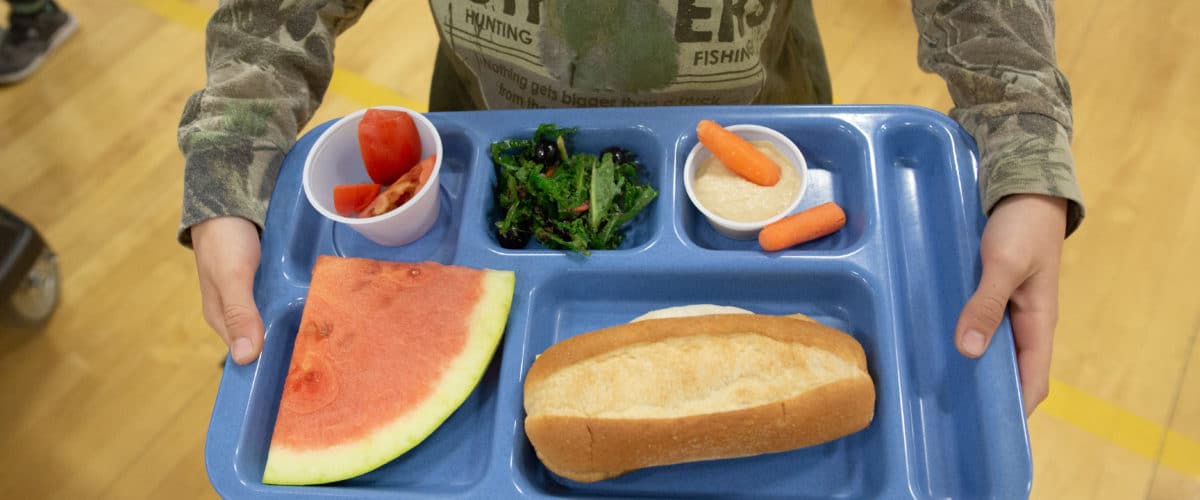 child holding a blue lunch tray with watermelon, carrots, greens, and tomatoes