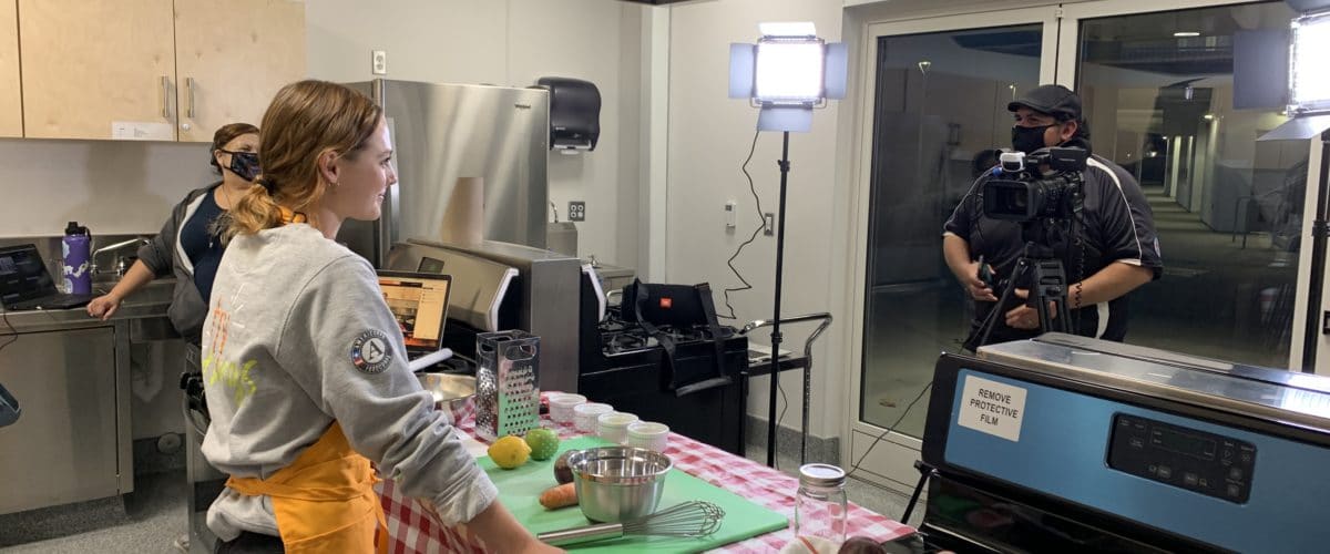 A FoodCorps service member wears a gold apron and stands in front of a table with a red checkered tablecloth. On the table is a green cutting board, a measuring bowl, a whisk and other kitchen tools. Across from the table, a person stands behind a camera on a tripod and a bright light shines in the corner.