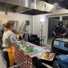 A FoodCorps service member wears a gold apron and stands in front of a table with a red checkered tablecloth. On the table is a green cutting board, a measuring bowl, a whisk and other kitchen tools. Across from the table, a person stands behind a camera on a tripod and a bright light shines in the corner.